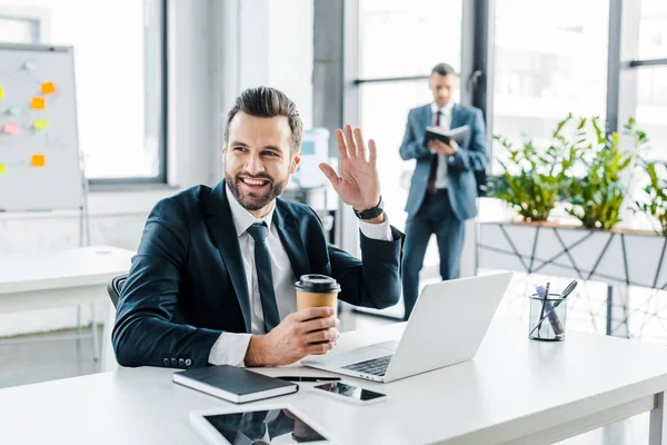 Selektiver Fokus eines gut gelaunten Mannes in formeller Kleidung, der mit einem Kollegen im Hintergrund winkt — Stockfoto