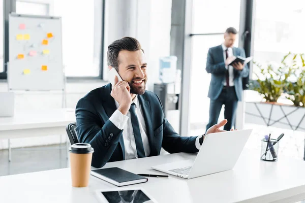 Focalizzazione selettiva di un uomo d'affari sorridente che parla su smartphone con un collega sullo sfondo — Foto stock