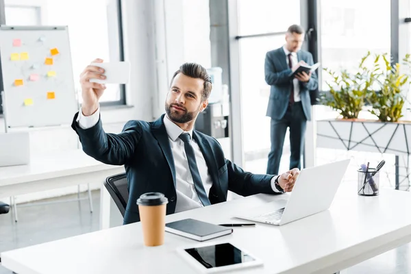 Messa a fuoco selettiva di felice uomo d'affari prendendo selfie in ufficio moderno — Foto stock