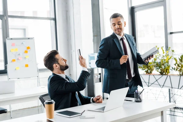 Foco selectivo de hombre de negocios guapo tomar una foto de compañero de trabajo alegre - foto de stock