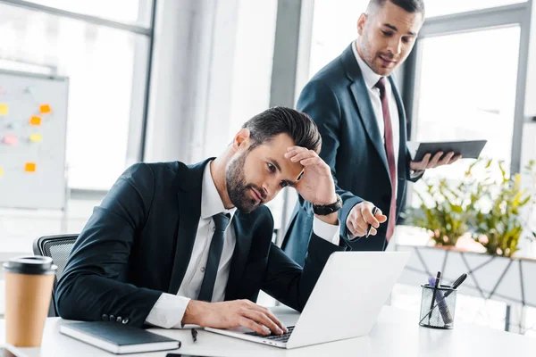 Enfoque selectivo de hombre de negocios guapo utilizando el ordenador portátil cerca de compañero de trabajo en la oficina moderna - foto de stock