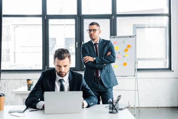 Selective focus of handsome businessman in glasses standing with crossed arms and looking at man typing on laptop — Stock Photo