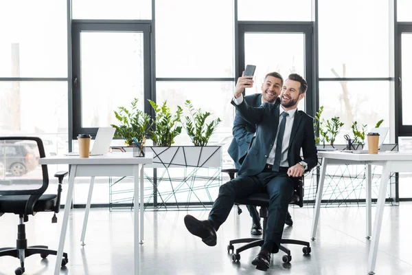 Busimessmen guapos tomando selfie y sonriendo en oficina moderna - foto de stock