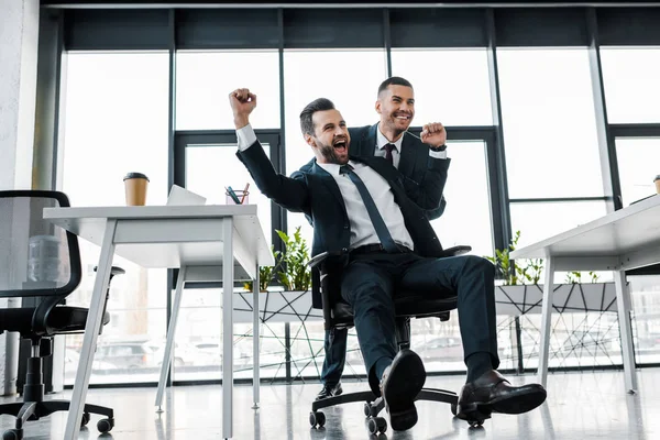 Homem de negócios feliz montando na cadeira perto do colega de trabalho no escritório moderno — Fotografia de Stock