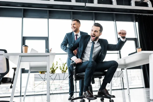 Homem de negócios alegre montando na cadeira perto do colega de trabalho no escritório moderno — Fotografia de Stock