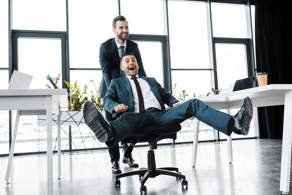 Homem de negócios feliz empurrando cadeira com colega de trabalho alegre no escritório moderno — Fotografia de Stock