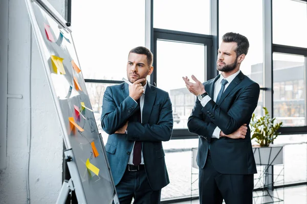 Hombres de negocios guapos mirando la pizarra blanca y hablando en la oficina moderna - foto de stock