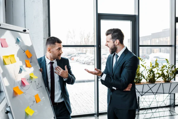 Homens de negócios irritados gritando com colega de trabalho perto de placa branca no escritório moderno — Fotografia de Stock