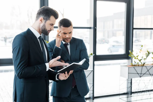 Handsome businessman looking at notebook near colleague in modern office — Stock Photo