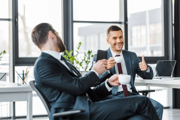 Selective focus of smiling businessman holding cup with drink and showing thumb up near colleague — Stock Photo