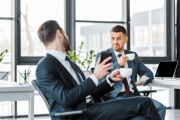 Enfoque selectivo de hombre de negocios sosteniendo taza con bebida y sentado cerca de colega - foto de stock