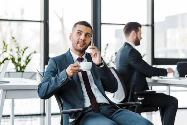 Enfoque selectivo de hombre de negocios sosteniendo la taza y hablando en el teléfono inteligente cerca de colega utilizando el ordenador portátil - foto de stock
