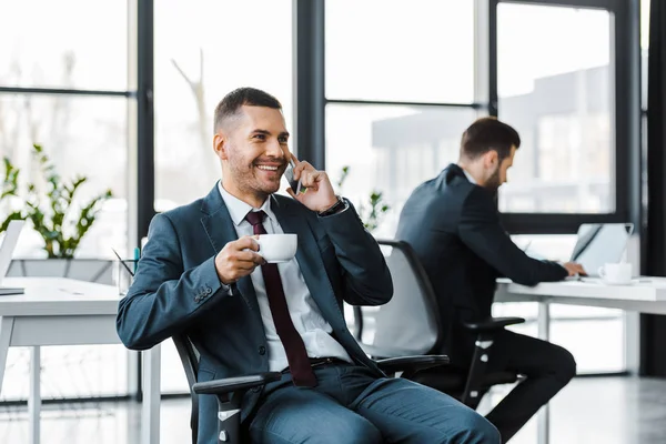 Selektiver Fokus des gut gelaunten Geschäftsmannes, der Tasse in der Hand hält und mit Laptop auf dem Smartphone neben dem Kollegen spricht — Stockfoto