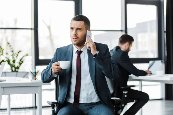 Selektiver Fokus des wütenden Geschäftsmannes, der Tasse mit Getränk in der Hand hält und in der Nähe des Kollegen mit dem Smartphone spricht — Stockfoto