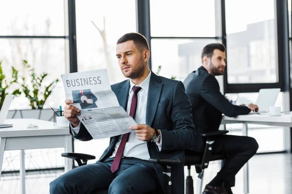 Selektiver Fokus eines gutaussehenden Geschäftsmannes beim Lesen einer Wirtschaftszeitung in der Nähe eines Mitarbeiters — Stockfoto
