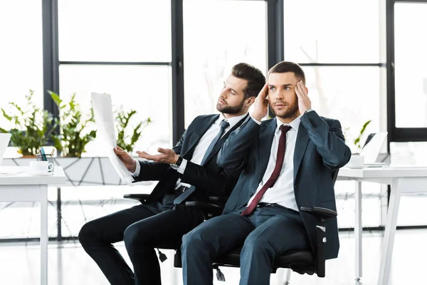 Handsome businessman reading newspaper near coworker holding head in modern office — Stock Photo