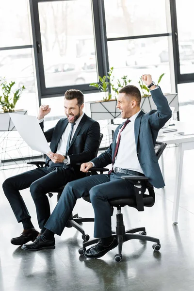 Heureux hommes d'affaires regardant le papier et célébrant la victoire dans le bureau moderne — Photo de stock