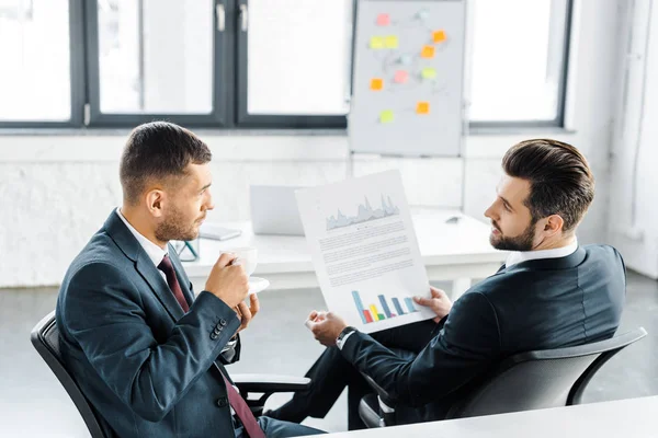 Enfoque selectivo del empresario sosteniendo papel con gráficos y hablando con su compañero de trabajo - foto de stock