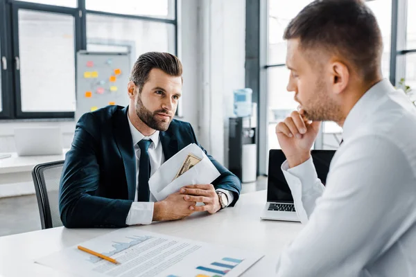 Hombre de negocios barbudo sosteniendo sobre con soborno cerca de compañero de trabajo en la oficina moderna - foto de stock