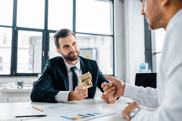 Selektiver Fokus des bärtigen Geschäftsmannes, der Dollar-Banknoten in der Nähe eines Mitarbeiters im modernen Büro hält — Stockfoto