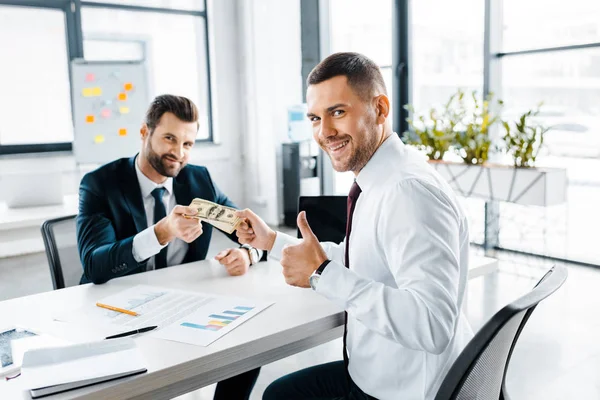 Cheerful businessman showing thumb up while taking dollar banknotes in modern office — Stock Photo