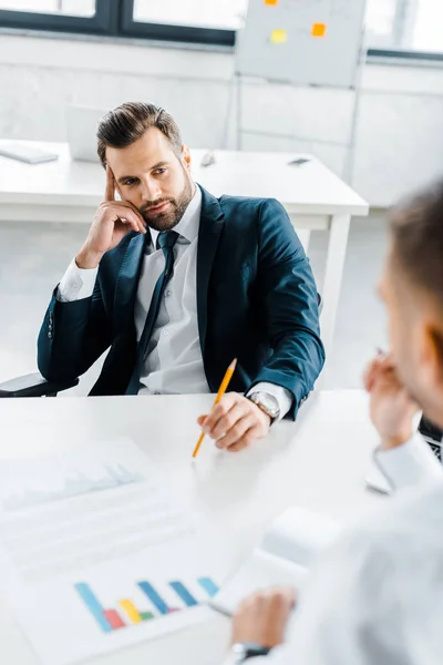 Uomo d'affari barbuto in possesso di matita e guardando collega mentre seduto alla scrivania — Foto stock