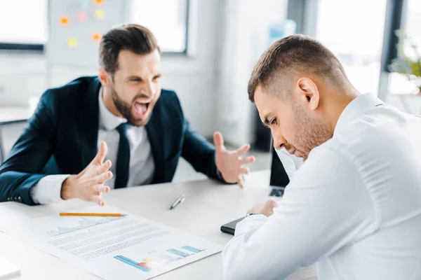 Selective focus of upset man sitting near angry man in formal wear screaming in modern office — Stock Photo