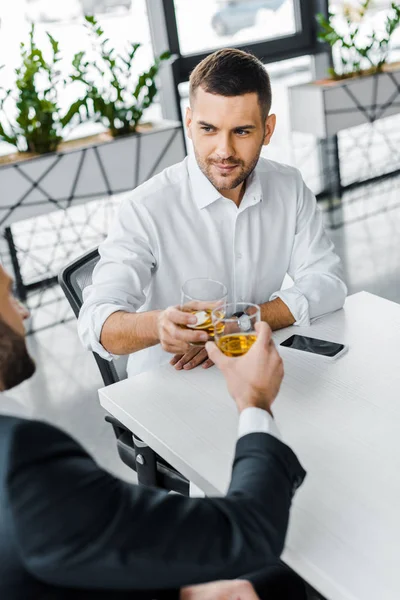 Guapo hombre de negocios tostando con whisky mientras está sentado en la oficina moderna con su compañero de trabajo - foto de stock