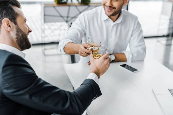 Barbudo hombre de negocios tostando con whisky mientras está sentado en la oficina moderna con su compañero de trabajo - foto de stock
