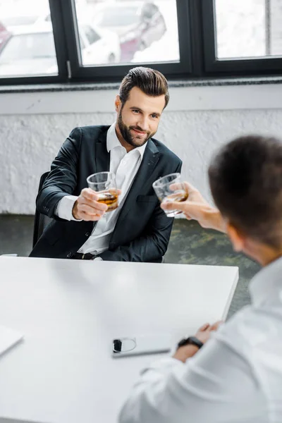 Barbudo hombre de negocios sosteniendo vidrio ow whisky mientras está sentado en la oficina moderna con su compañero de trabajo - foto de stock