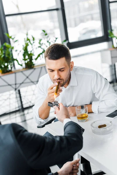 Empresário em uso formal fumar charuto cubano perto de colega de trabalho segurando isqueiro no escritório moderno — Fotografia de Stock