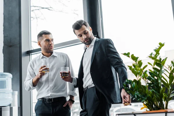 Uomo d'affari in piedi con la mano in tasca e tenendo un bicchiere di whisky vicino al collega con sigaro cubano — Foto stock