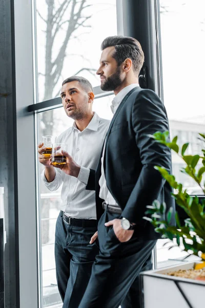 Bearded businessman standing with hand in pocket and holding glass of whiskey near coworker — Stock Photo