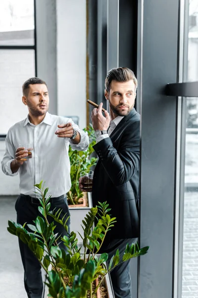 Businessman pointing with finger while holding glass of whiskey near coworker with cuban cigar — Stock Photo