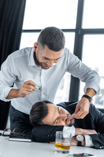 Homem alegre que prende o alfinete perto do nariz do colega dormindo no escritório moderno — Fotografia de Stock