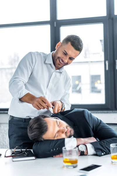 Uomo allegro che punta con pennarello sul volto del collega addormentato in ufficio moderno — Foto stock