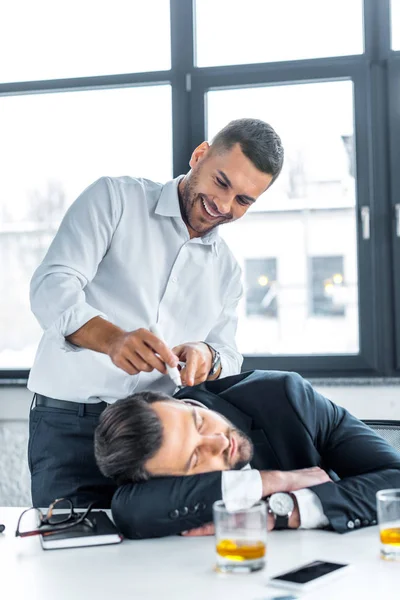 Hombre alegre señalando con marcador en la cara de compañero de trabajo durmiendo en la oficina moderna - foto de stock