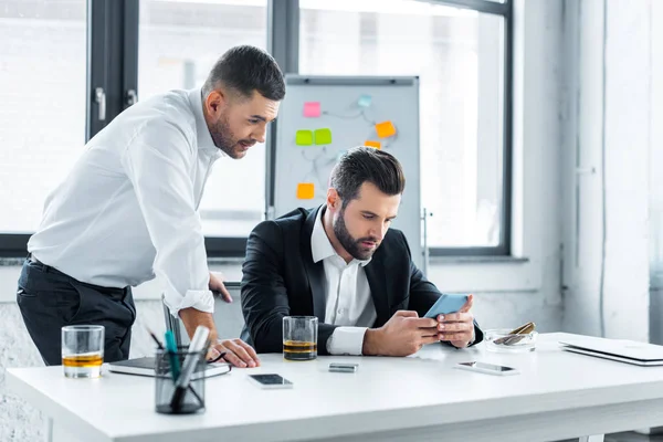 Hombre de negocios beadred celebración de teléfono inteligente cerca de compañero de trabajo en la oficina - foto de stock