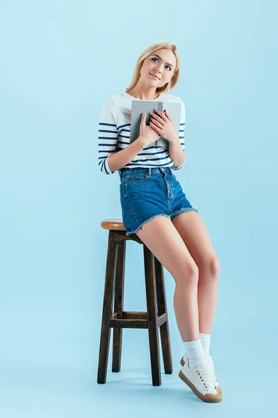 Мечтательная девушка с книгой, сидящей на деревянном стуле на синем фоне — стоковое фото