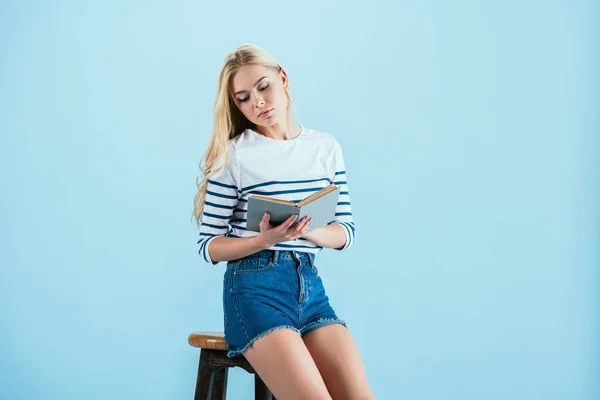 Encantadora mujer sentada en la silla y leyendo libro aislado en azul - foto de stock