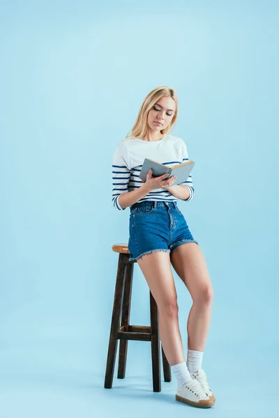 Maravilhosa jovem sentada na cadeira e lendo livro sobre fundo azul — Fotografia de Stock