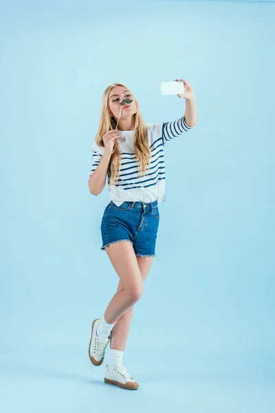 Fille blonde avec fausse moustache prendre selfie sur fond bleu — Photo de stock