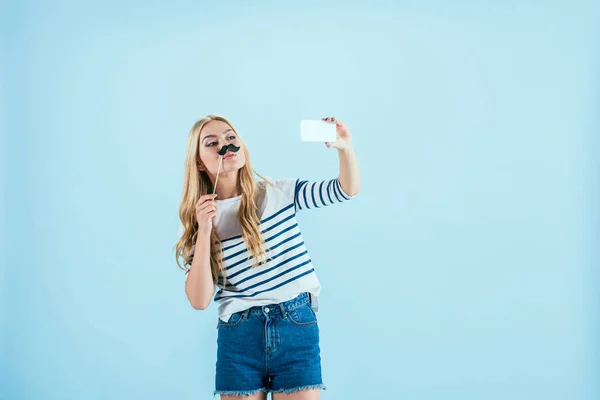 Attractive girl holding fake mustache and taking selfie on blue background — Stock Photo