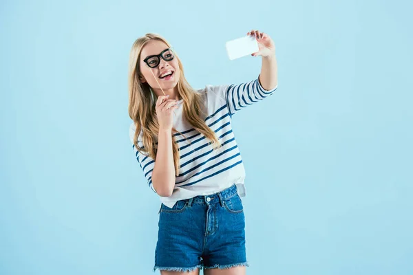 Lachendes Mädchen mit Spielzeugbrille macht Selfie auf blauem Hintergrund — Stockfoto