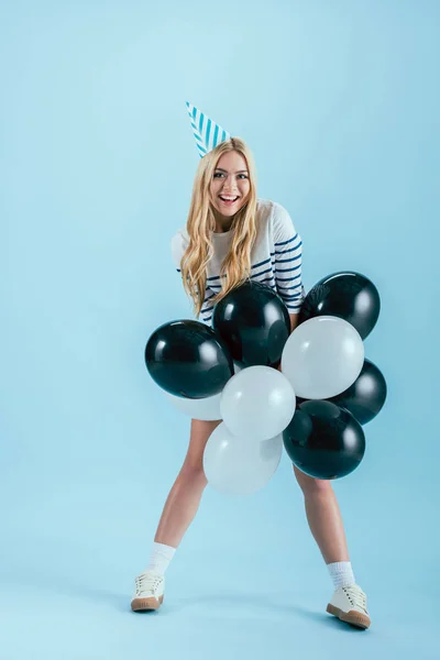 Смешная блондинка в шляпе для вечеринок позирует с воздушными шарами на синем фоне — стоковое фото