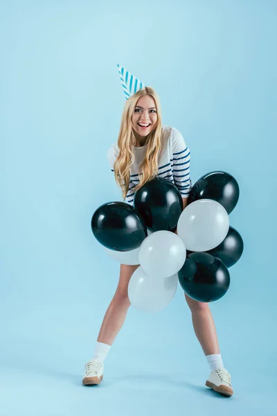 Divertente ragazza bionda in cappello partito in posa con palloncini su sfondo blu — Foto stock