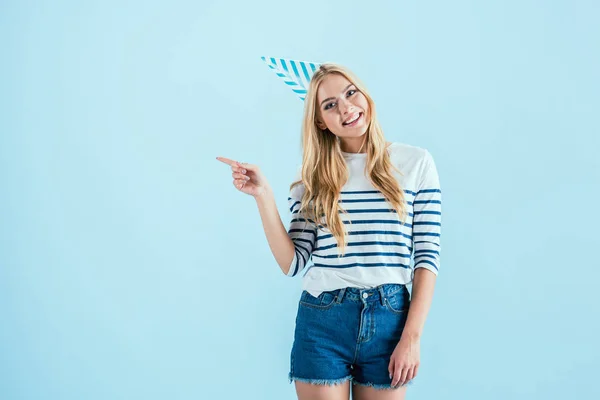 Studio shot de fille blonde en chapeau de fête pointant avec le doigt isolé sur bleu — Photo de stock