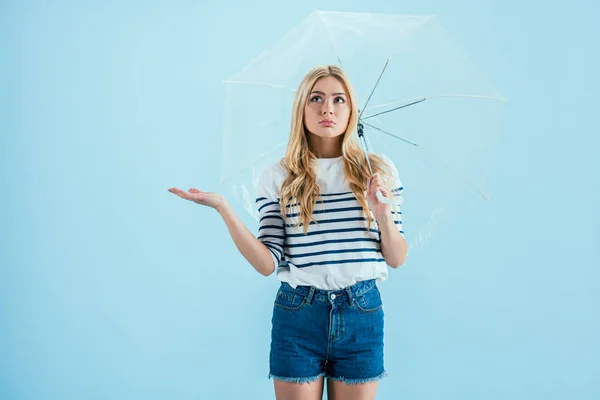 Molesto chica rubia de pie bajo el paraguas sobre fondo azul - foto de stock