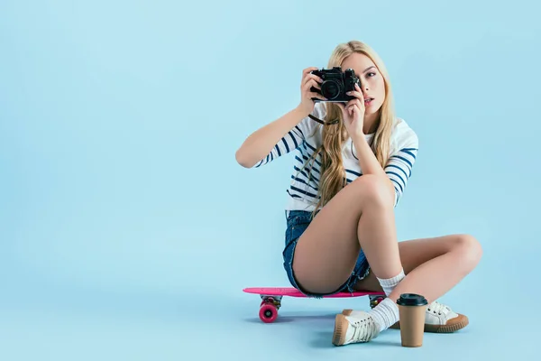 Studio shot of girl sitting on longboard and taking picture on blue background — Stock Photo