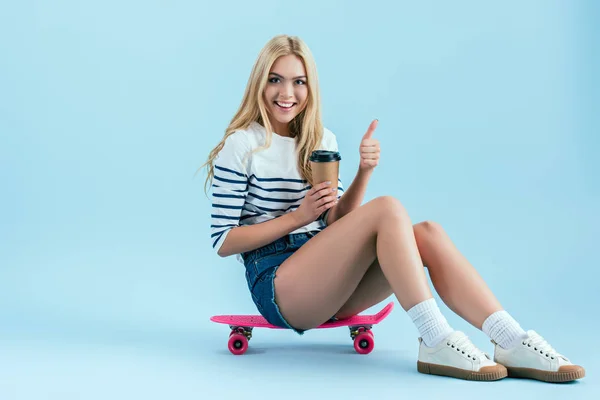 Cheerful girl with cup of coffee sitting on longboard and showing thumb up on blue background — Stock Photo